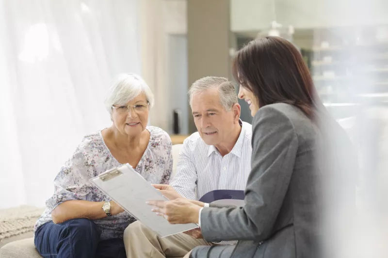 An elderly couple look hopeful as they consult with a female physician holding a clipboard.